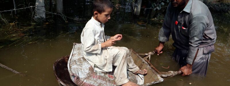Ein Mann schiebt seine Schubkarre, in der ein Junge sitzt, durch das Hochwasser. - Foto: Muhammad Sajjad/AP/dpa