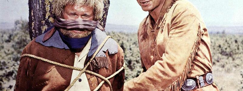Old Shatterhand (Lex Barker, r) hilft dem neugierigen Sam Hawkins (Ralf Wolter, l), von einem Marterpfahl loszukommen (1965). - Foto: United Archives/IFTN/dpa