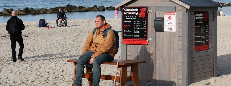 Ein Mann sitzt am Ostseestrand vor dem Häuschen eines Strandkorbvermieters und genießt das warme und sonnige Wetter. - Foto: Bernd Wüstneck/dpa