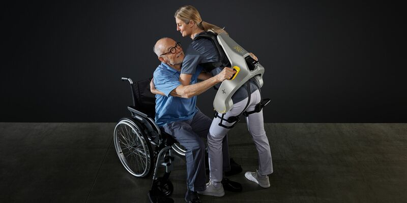 Eine Frau hebt mit Hilfe eines intelligenten Kraftanzugs der Firma German Bionic einen Mann aus einem Rollstuhl. - Foto: German Bionic/dpa