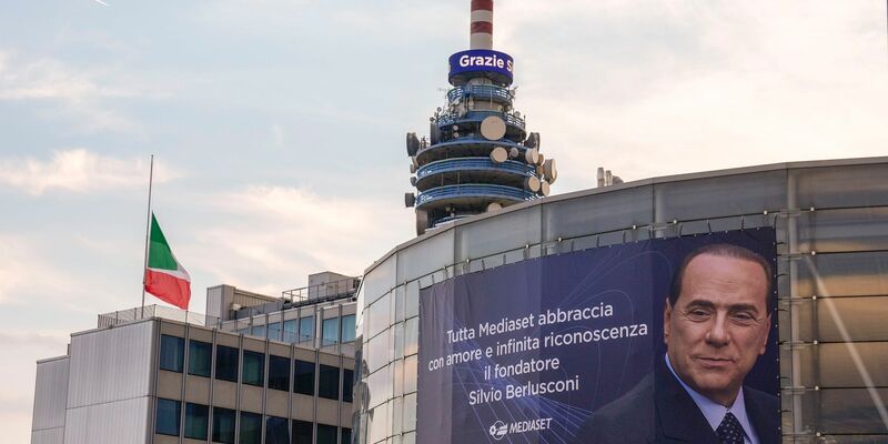 Eine italienische Flagge auf Halbmast weht vor einer Nachricht mit der Aufschrift «Grazie Silvio» auf einem Turm des Fernsehsenders Mediaset neben einem großen Plakat. - Foto: Luca Bruno/AP