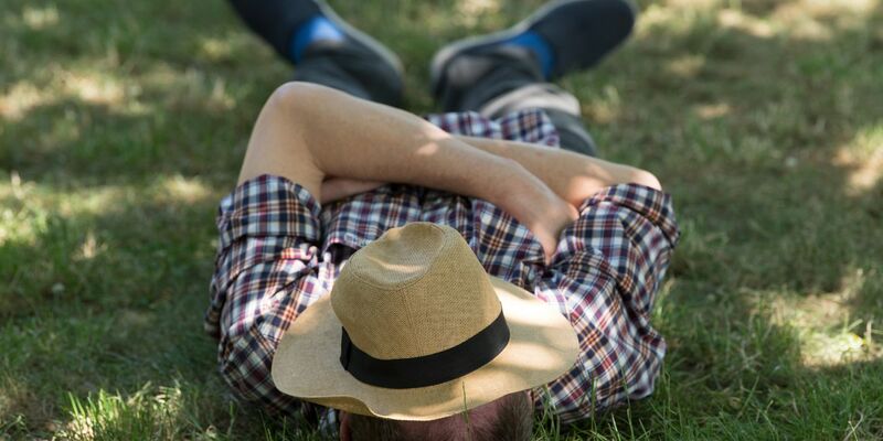 Ein Mann mit einem Sonnenhut auf dem Gesicht liegt auf einer Wiese im Schatten und ruht sich aus. - Foto: Sebastian Kahnert/zb/dpa