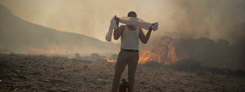 Ein Mann schützt sich mit einem Handtuch vor dem Rauch, während er versucht, ein Feuer nahe des Badeortes Lindos zu löschen. - Foto: Petros Giannakouris/AP/dpa