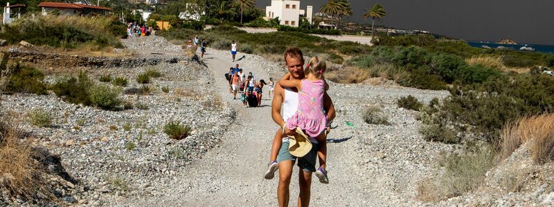 Ein Mann mit einem Kind auf dem Arm verlässt ein Waldbrandgebiet auf Rhodos. - Foto: Lefteris Damianidis/InTime News/AP/dpa