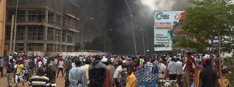 Mit dem brennenden Hauptquartier der Regierungspartei im Rücken demonstrieren Anhänger meuternder Soldaten in der Hauptstadt Niamey. - Foto: Fatahoulaye Hassane Midou/AP/dpa