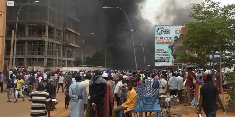 Mit dem brennenden Hauptquartier der Regierungspartei im Rücken demonstrieren Anhänger meuternder Soldaten. - Foto: Fatahoulaye Hassane Midou/AP/dpa