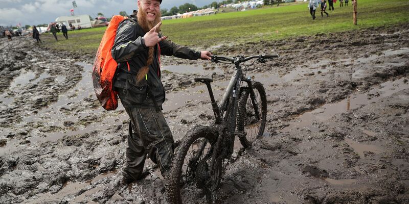 Metal-Fan Lukas Litwin aus Bochum ist mit seinem E-Mountainbike auf dem schlammigen Festivalgelände unterwegs. Noch ist unklar, wie viele der erwarteten 85.000 Metalfans tatsächlich auf Wackens Ackern feiern. - Foto: Christian Charisius/dpa