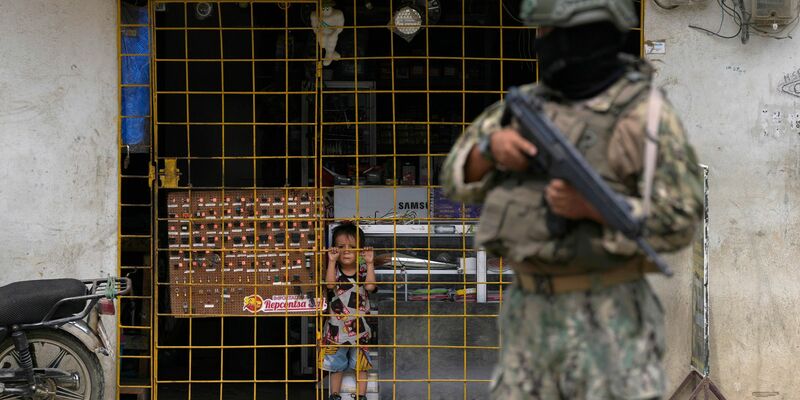 Ein Junge schaut hinter den Gitterstäben eines geöffneten Elektronikgeschäfts hervor, während ein Soldat an einem von der Armee eingerichteten Sicherheitskontrollpunkt Wache steht. - Foto: Martin Mejia/AP/dpa