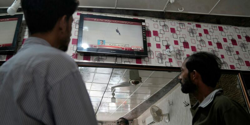 Menschen in einem Friseur-Salon verfolgen eine Nachrichtensendung, in der über eine riskante Rettungsmission an der Seilbahn berichtet wird. - Foto: K.M. Chaudary/AP/dpa