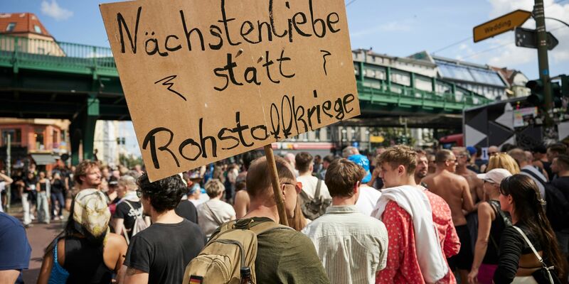 Ein Feiernder trägt ein Schild mit der Aufschrift «Nächstenliebe statt Rohstoffkriege» bei der Technoparade Zug der Liebe durch Berlin. - Foto: Joerg Carstensen/dpa