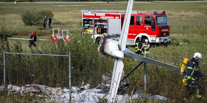 Trümmerteile eines Flugzeugs hängen in einem Zaun, während Einsatzkräfte der Feuerwehr an der Absturzstelle stehen. - Foto: Pia Bayer/dpa