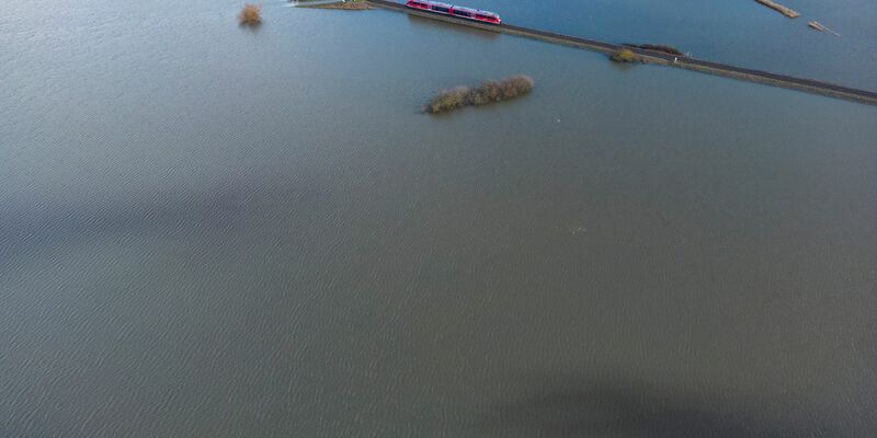 Ein Personenzug der Deutschen Bahn fährt im Januar 2023 durch eine überschwemmte Flussniederung bei Eichen in Hessen. - Foto: Boris Roessler/dpa