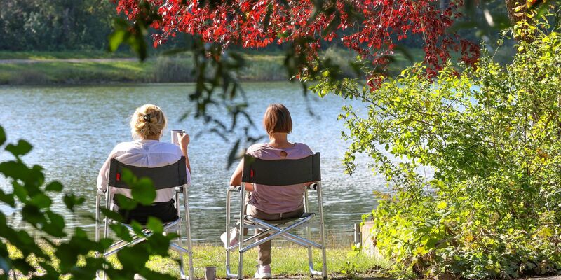 Zwei Frauen sitzen im baden-württembergischen Ertingen an einem See in der Sonne. - Foto: Thomas Warnack/dpa