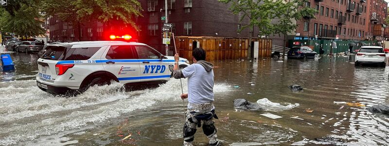 Hochwasser auf den Straßen von New York: Im Stadtteil Brooklyn versucht ein Mann, mit einem  Stock einen Abfluss zu reinigen, während ein Polizeiwagen vorbeifährt. - Foto: Jake Offenhartz/AP/dpa