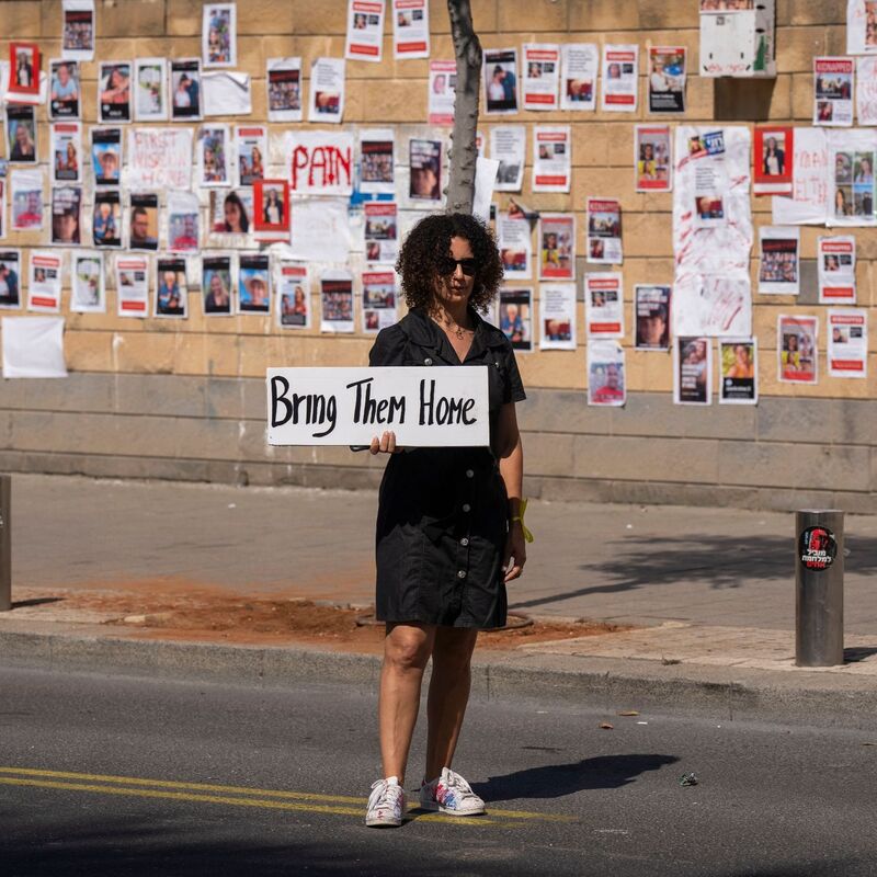 «Bringt sie nacht Hause» steht auf dem Schild dieser Frau, die in Tel Aviv vor einer Wand mit den Fotos von Israelis steht, die von der Hamas im Gazastreifen gefangen gehalten werden. - Foto: Petros Giannakouris/AP/dpa