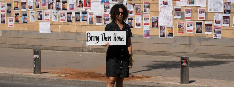 «Bringt sie nacht Hause» steht auf dem Schild dieser Frau, die in Tel Aviv vor einer Wand mit den Fotos von Israelis steht, die von der Hamas im Gazastreifen gefangen gehalten werden. - Foto: Petros Giannakouris/AP/dpa