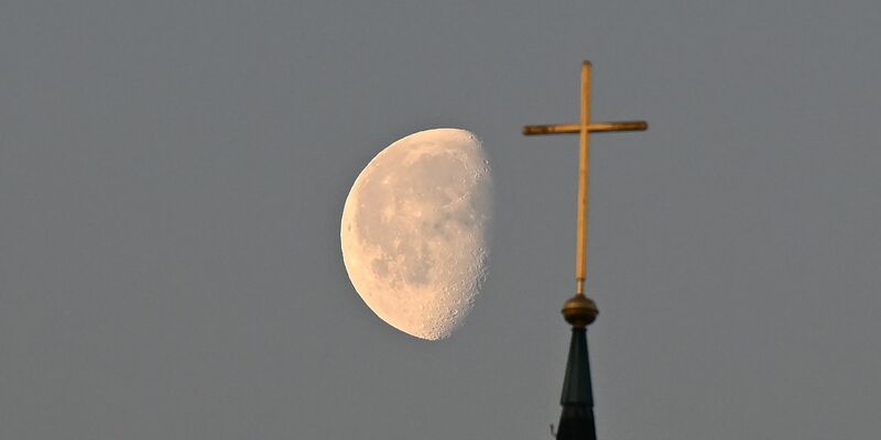 Der Mond im Morgenlicht hinter einem Kreuz auf einem Kirchturm. - Foto: Bernd Weißbrod/dpa