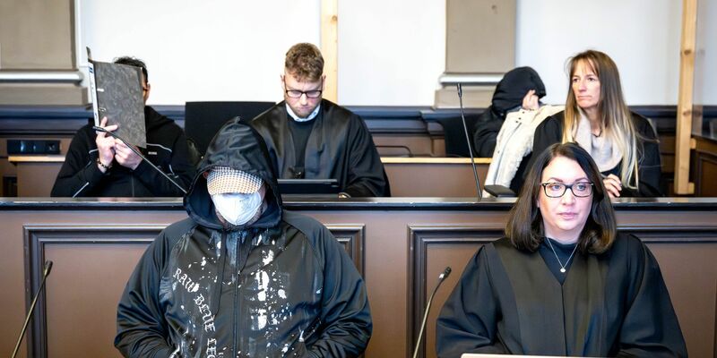 Die drei Angeklagten (l, vorne l sowie hinten r) sitzen vor Prozessbeginn mit ihren Anwältinnen und Anwälten im Gerichtssaal. - Foto: Sina Schuldt/dpa