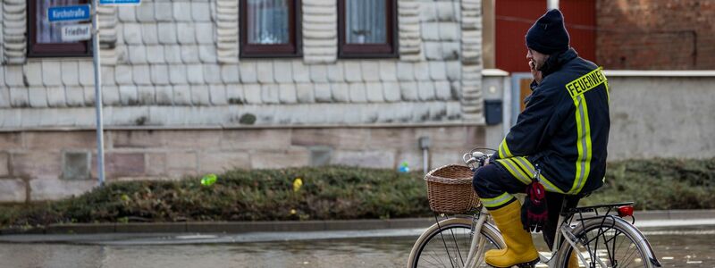 Ein Feuerwehrmann auf einem Fahrrad in einer überschwemmten Straße in Windehausen. - Foto: Christoph Reichwein/dpa