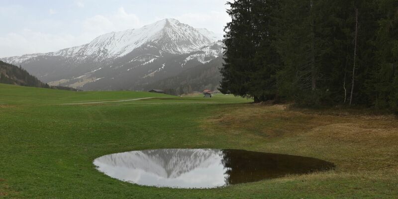 Die im Dunst liegenden Alpen spiegeln sich in einem Weiher. Der Saharastaub-Höhepunkt ist überschritten. - Foto: Karl-Josef Hildenbrand/dpa