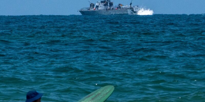 Ein Surfer wartet auf eine Welle, während ein israelisches Marineschiff im Mittelmeer vor der Küste von Hadera patrouilliert. - Foto: Ariel Schalit/AP
