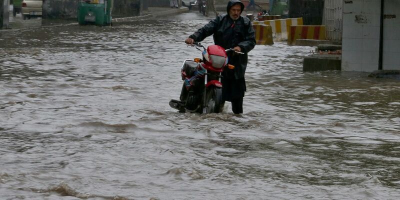 Ein Pakistaner watet nach starken Regenfällen mit seinem Motorrad durch eine überschwemmte Straße in Peschawar. - Foto: Muhammad Sajjad/AP/dpa