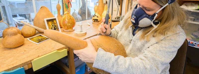 Virginia Lauterbach bearbeitet in ihrer Werkstatt einen Flaschenkürbis mit einem Bohrer. - Foto: Julian Stratenschulte/dpa