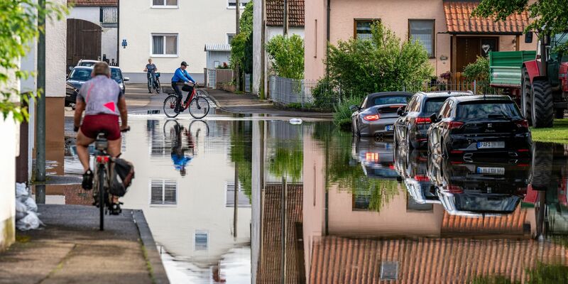 Radler fahren durch eine überflutete Straße. Im Bereich der Gemeinde Baar-Ebenhausen war ein Damm an zwei Stellen geborsten. - Foto: Armin Weigel/dpa