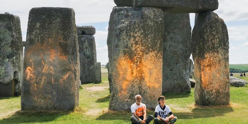 Demonstranten von «Just Stop Oil» sitzen vor dem Steinmonument Stonehenge, das sie zuvor mit orangener Farbe besprüht haben. - Foto: Just Stop Oil/PA Media/dpa