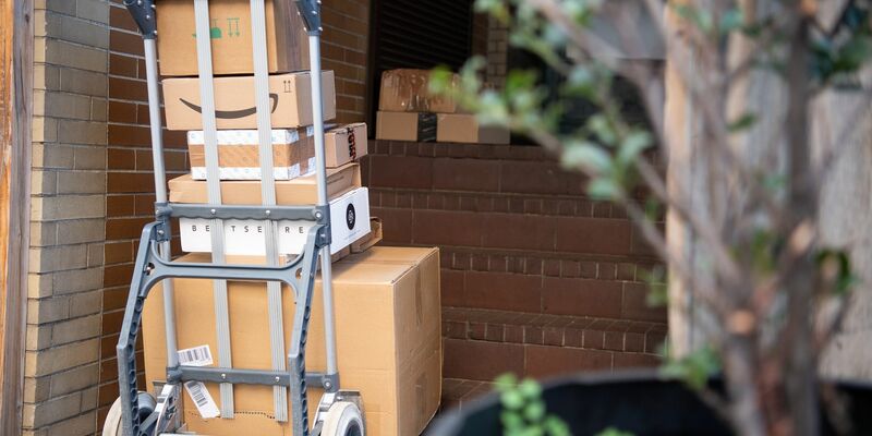 Pakete stehen auf einer Karre, während ein Paketzusteller Pakete ausliefert. - Foto: Sebastian Gollnow/dpa