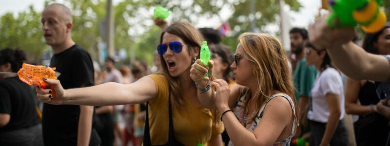 Ein Mann und eine Frau auf der Terrasse eines Restaurants in Barcelona lassen sich von vorbeiziehenden Demonstranten gegen den Massentourismus offenbar nicht aus der Ruhe bringen. - Foto: Lorena Sopêna/EUROPA PRESS/dpa