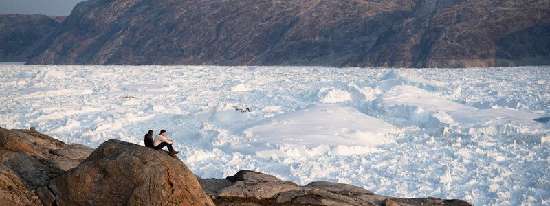 Zwei Studenten der New York University sitzen auf einem Felsen mit Blick auf den grönländischen Helheim-Gletscher.  - Foto: Felipe Dana/AP/dpa